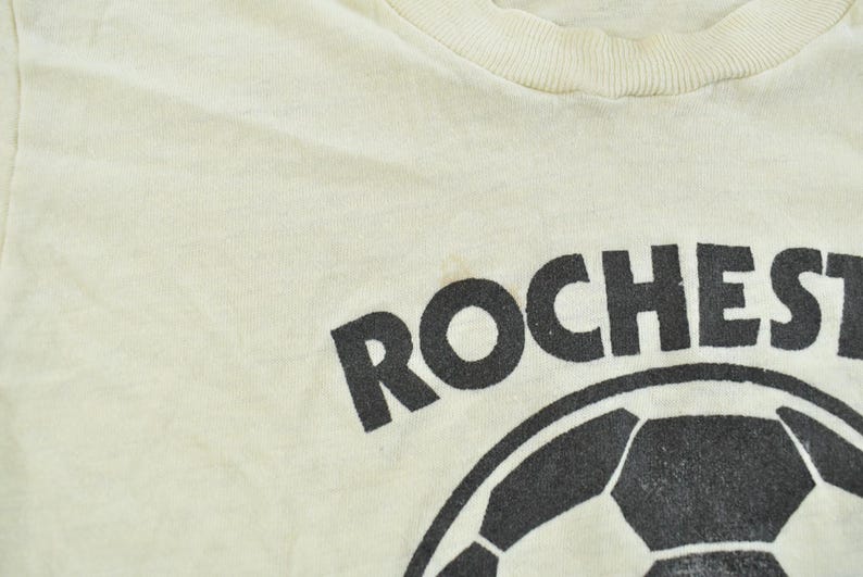 May include: A cream-colored t-shirt featuring a black graphic print of a soccer ball and the partial text ROCHEST in a bold, sans-serif font. The garment has a ribbed crew neckline and a soft, lightweight fabric construction.
