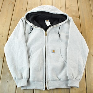 Puede incluir: Sudadera con capucha y cremallera gris claro con capucha forrada en negro. La sudadera tiene un bolsillo delantero a cada lado y un peque&ntilde;o parche con el logotipo en el pecho izquierdo. La cremallera es dorada.