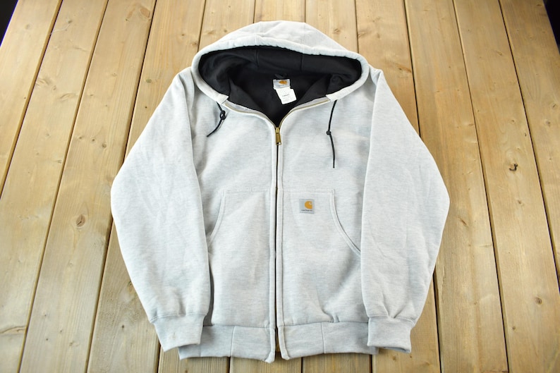 Puede incluir: Sudadera con capucha y cremallera gris claro con capucha forrada en negro. La sudadera tiene un bolsillo delantero a cada lado y un peque&ntilde;o parche con el logotipo en el pecho izquierdo. La cremallera es dorada.