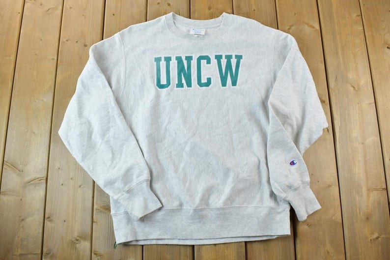 Puede incluir: Una sudadera gris con el texto "UNCW" en letras verde azulado en la parte delantera. La sudadera tiene un logotipo de Champion en la manga izquierda.