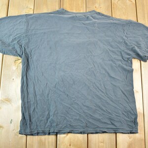 May include: A faded blue-grey t-shirt, laid flat on a wooden surface. The shirt has short sleeves and a crew neck. The fabric is wrinkled, suggesting it has been worn and washed.