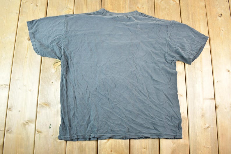 May include: A faded blue-grey t-shirt, laid flat on a wooden surface. The shirt has short sleeves and a crew neck. The fabric is wrinkled, suggesting it has been worn and washed.
