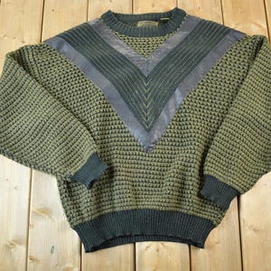 Könnte beinhalten: Olivgrüner Pullover mit strukturiertem Strickmuster und einem dunkelgrauen V-förmigen Design auf der Brust. Der Pullover hat gerippte Bündchen und Saum. Die Ärmel sind lang und der Ausschnitt ist rund. Der Pullover ist aus weichem Material.