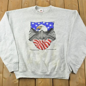 May include: Grey sweatshirt featuring a graphic of a bald eagle flying over a fighter jet with an American flag in the background. The text "Whiteman A.F.B. Knob Noster, MO" is printed below the graphic.