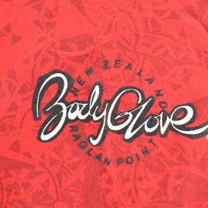 May include: Red fabric with a repeating geometric pattern. The words "Body Glove" are in a white script font with a black outline. Below the text is "New Zealand Raglan Point" in a circular arrangement.