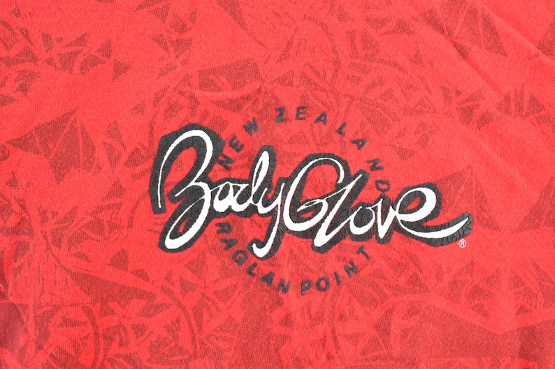 May include: Red fabric with a repeating geometric pattern. The words "Body Glove" are in a white script font with a black outline. Below the text is "New Zealand Raglan Point" in a circular arrangement.