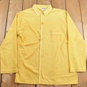 May include: A vintage yellow button-down shirt with long sleeves and a chest pocket. The shirt has a collared neckline and is made of a lightweight fabric. The shirt is laid flat on a wooden surface.