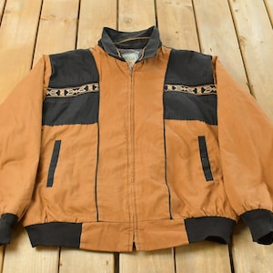 May include: A vintage brown and black jacket with a full zip. The jacket has black panels on the shoulders and cuffs, with decorative patterns. It features two front pockets and a stand-up collar.
