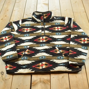 May include: A fleece pullover featuring a high collar with a snap button placket and a front kangaroo pocket. The garment displays a repeating geometric pattern in black, cream, red, and blue, with long sleeves and a straight hemline finish.
