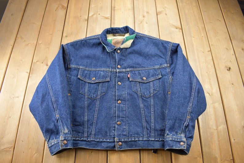 May include: A blue denim jacket with a brown button closure and a plaid flannel lining. The jacket has two front pockets and a single pocket on the left chest.