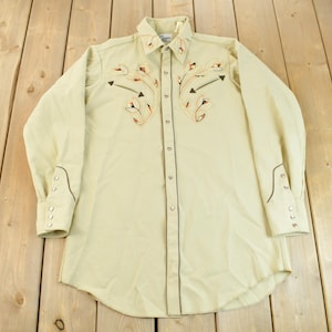 May include: A light beige long-sleeved shirt with a western style embroidered design on the front. The embroidery features brown and orange floral patterns with arrows.