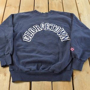 Puede incluir: Sudadera azul marino de Georgetown con letras blancas arqueadas. La sudadera tiene cuello redondo, mangas largas y un pequeño emblema en la manga derecha. La tela parece ser un material suave y desgastado.