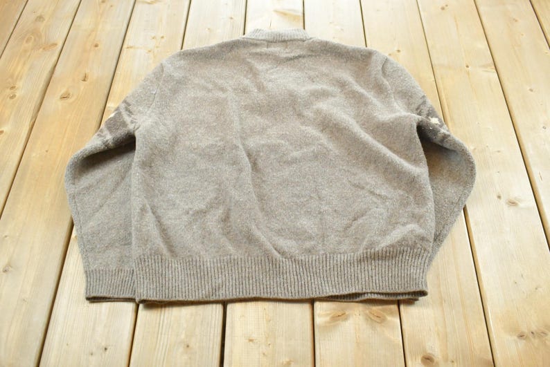 Peut inclure: Un pull &agrave; manches longues marron clair avec un ourlet et des poignets c&ocirc;tel&eacute;s. Le dos du pull est visible, montrant la texture du tissu tricot&eacute;. Le pull a un col rond et un design simple et classique.