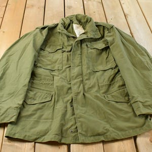 May include: An olive green field jacket featuring a high collar, a front zipper closure with a snap-button storm flap, and four large rectangular pockets on the front. The garment includes long sleeves with adjustable cuffs and a white interior label.