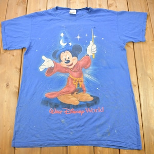 Pu&ograve; includere: Una maglietta blu con un'immagine sbiadita di Topolino vestito da mago, che tiene una bacchetta magica. La maglietta ha la scritta "Walt Disney World" stampata in basso.