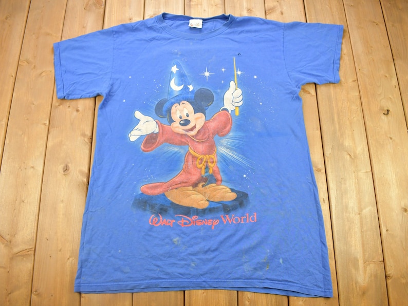 Pu&ograve; includere: Una maglietta blu con un'immagine sbiadita di Topolino vestito da mago, che tiene una bacchetta magica. La maglietta ha la scritta "Walt Disney World" stampata in basso.