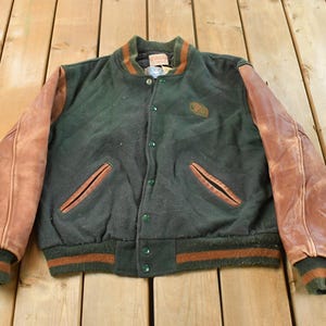 Könnte beinhalten: Eine Vintage-Collegejacke in Dunkelgrün und Braun. Die Jacke hat braune Lederärmel, einen grünen Körper und einen grün-braun gestreiften Kragen, Bündchen und Saum. Sie hat eine Knopfleiste und zwei Vordertaschen.