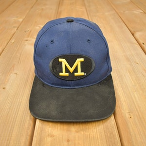 Op de afbeelding: Een blauwe en zwarte baseballpet met een gele "M" geborduurd op de voorkant.