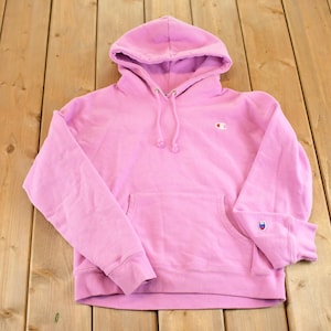 May include: A light pink Champion hoodie with a drawstring hood and a kangaroo pocket. The Champion logo is visible on the left sleeve and chest. The hoodie is made of a soft, comfortable material.