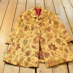 May include: A long-sleeved coat featuring a golden yellow base with a multi-colored floral and vine tapestry pattern in shades of red, green, and brown. The coat has a notched collar, a button-front closure, and a solid red interior lining.