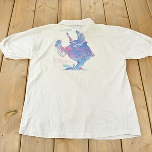 Può includere: Polo bianco con una stampa grafica colorata di una creatura con una persona che cavalca sulla sua schiena. La grafica è sul retro della maglietta.