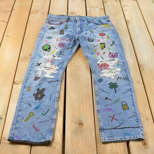 Puede incluir: Vaqueros de mezclilla azul claro con una variedad de ilustraciones coloridas dibujadas a mano, que incluyen un alienígena verde, un oso de peluche, un trébol, un candado y las palabras LOVE y MAN. Los vaqueros tienen zonas desgastadas con hilos blancos deshilachados.