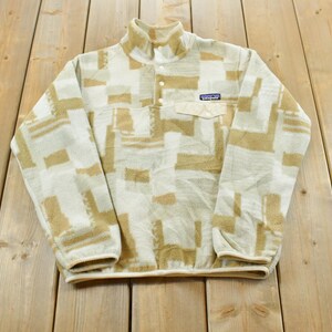 May include: A Patagonia fleece pullover featuring a cream and tan geometric block pattern. The garment has a high collar with a snap-button placket, long sleeves with elastic cuffs, and a single chest pocket with a flap and the Patagonia logo label.