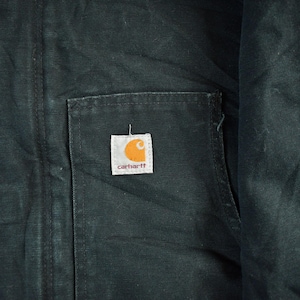 Puede incluir: Primer plano de una chaqueta Carhartt verde oscuro. La chaqueta presenta un bolsillo cuadrado con el logotipo de Carhartt, un cuadrado blanco con una 'C' naranja y el nombre de la marca en marr&oacute;n. La tela tiene una apariencia texturizada.