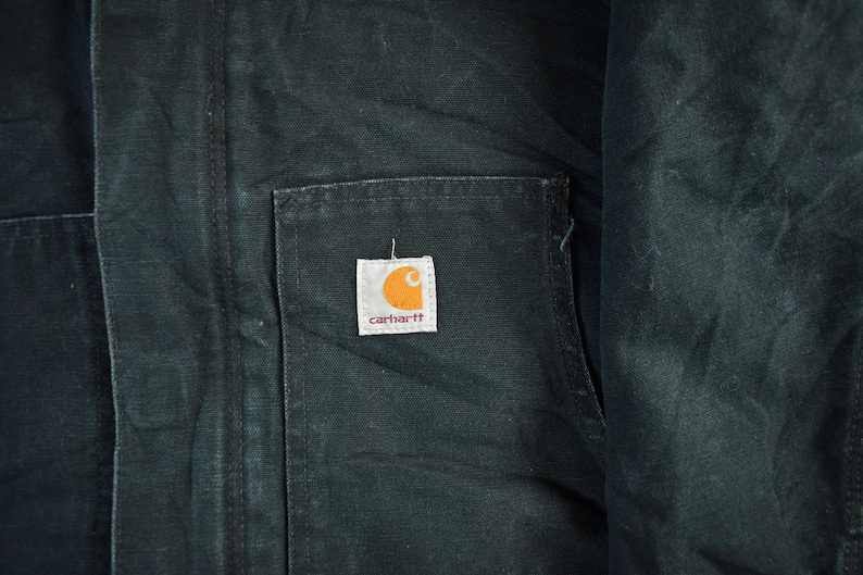 Puede incluir: Primer plano de una chaqueta Carhartt verde oscuro. La chaqueta presenta un bolsillo cuadrado con el logotipo de Carhartt, un cuadrado blanco con una 'C' naranja y el nombre de la marca en marr&oacute;n. La tela tiene una apariencia texturizada.