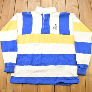 Può includere: Una maglia da rugby a maniche lunghe a righe bianche, blu e gialle con una piccola immagine ricamata di una persona a cavallo. La maglia ha un colletto abbottonato e una patta con bottoni.