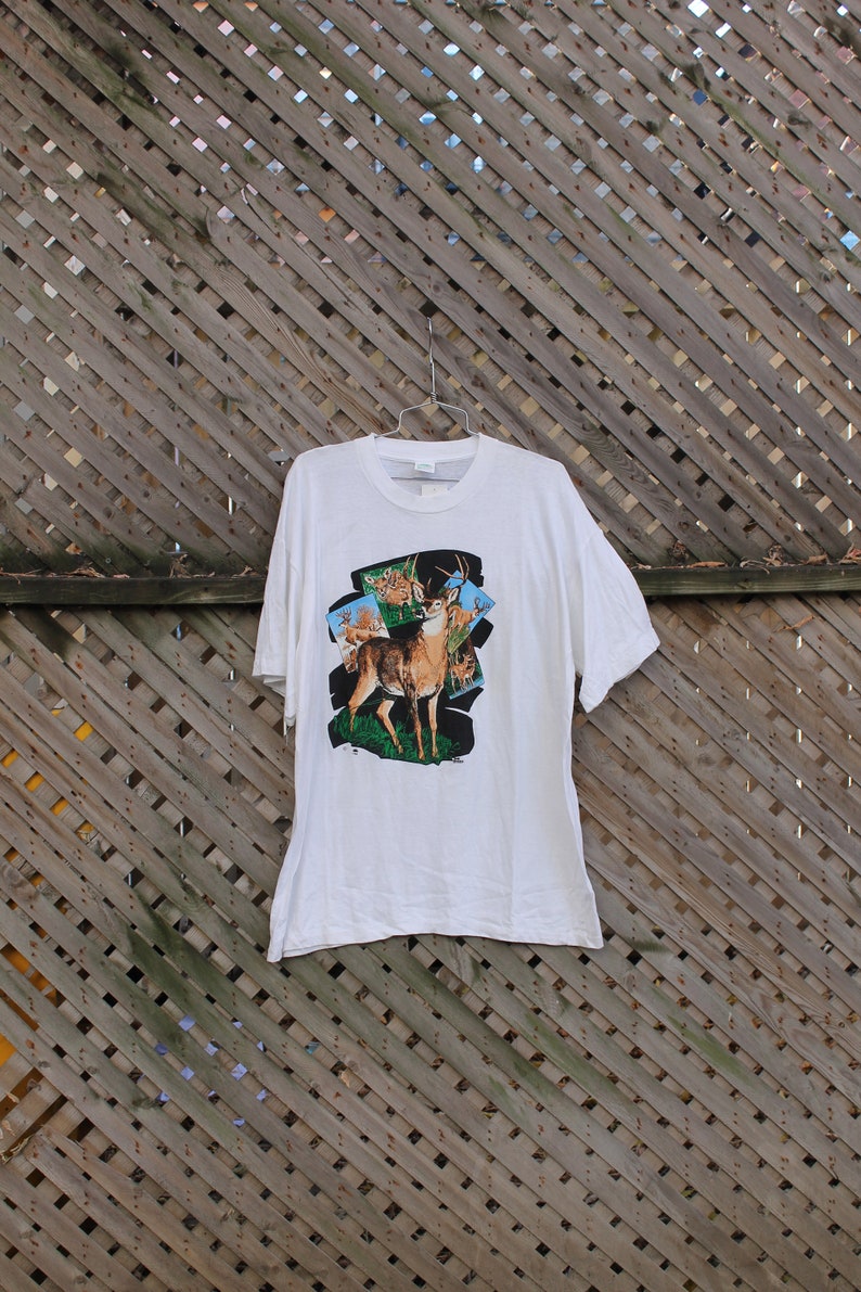 Pode incluir: Camiseta branca com uma estampa gr&aacute;fica de um veado com chifres em um ambiente de floresta. O veado &eacute; cercado por veados menores no fundo.