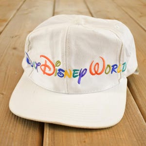 Può includere: Un cappellino da baseball bianco con la scritta "Disney World" ricamata in un arcobaleno di colori. Il cappellino ha una visiera curva e una corona strutturata, perfetto per una giornata al parco a tema.