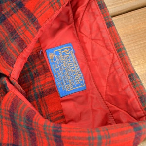 Puede incluir: Una camisa de franela a cuadros rojos y verdes con una etiqueta azul que dice "WARRANTED TO BE A PENDLETON TRADEMARK REG. U.S. PAT. OFF. PENDLETON WOOLEN MILLS PORTLAND, OREGON 100% PURE VIRGIN WOOL MADE IN USA"