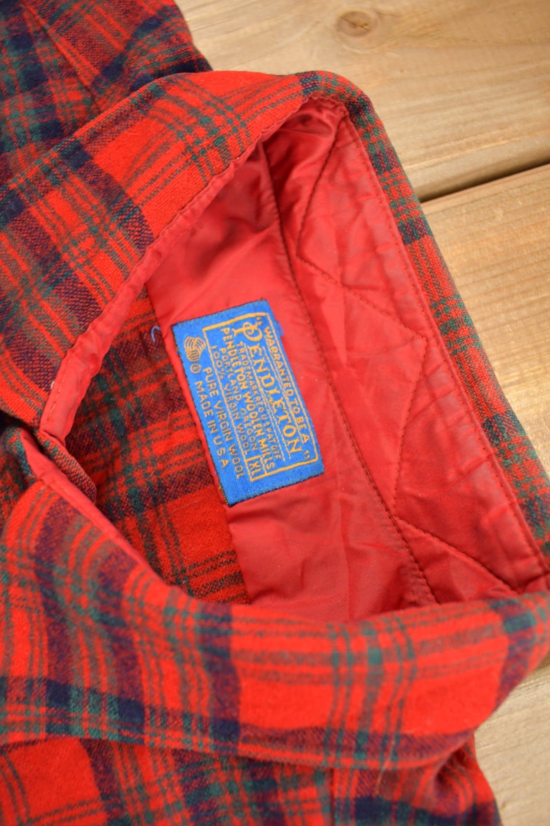 Puede incluir: Una camisa de franela a cuadros rojos y verdes con una etiqueta azul que dice "WARRANTED TO BE A PENDLETON TRADEMARK REG. U.S. PAT. OFF. PENDLETON WOOLEN MILLS PORTLAND, OREGON 100% PURE VIRGIN WOOL MADE IN USA"