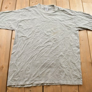 Könnte beinhalten: Ein hellgraues T-Shirt mit kurzen Ärmeln, flach auf einer Holzoberfläche liegend. Das Shirt hat einen Rundhalsausschnitt und scheint aus einem weichen, abgenutzten Stoff zu sein. Das Shirt ist leicht zerknittert, was darauf hindeutet, dass es getragen wurde.