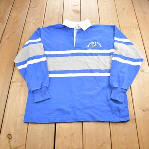 May include: A blue long-sleeved rugby shirt with white and gray stripes. The shirt has a white collar and a button placket. There is a small embroidered logo on the left chest.