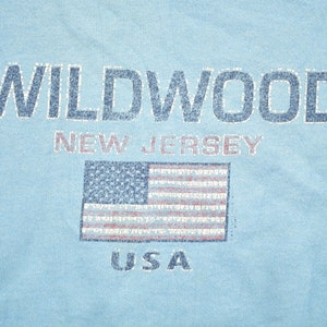 May include: A light blue t-shirt with the text "WILDWOOD NEW JERSEY USA" and a distressed American flag graphic.