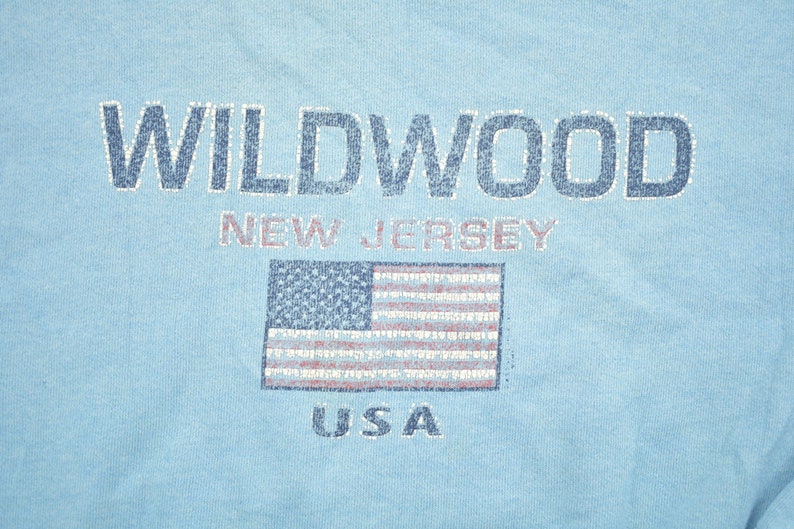 May include: A light blue t-shirt with the text "WILDWOOD NEW JERSEY USA" and a distressed American flag graphic.