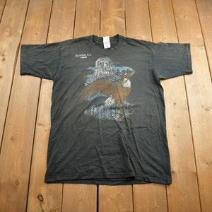May include: A dark grey t-shirt with a graphic print of an owl and an eagle. The text "BRITISH C.C. CANADA" is printed on the shirt.