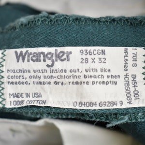 May include: A white tag with black text that reads "Wrangler" and includes care instructions. The tag is attached to a dark green fabric.