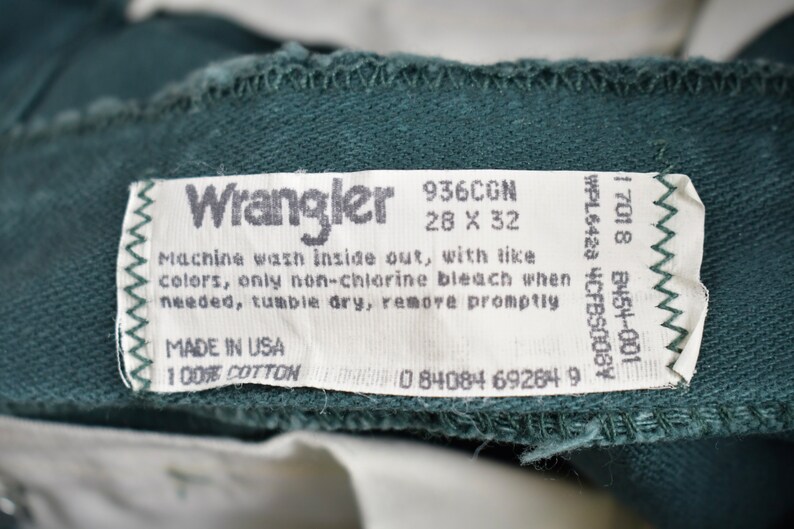 May include: A white tag with black text that reads "Wrangler" and includes care instructions. The tag is attached to a dark green fabric.