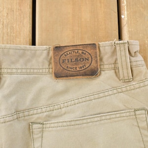 May include: Close-up of khaki-colored pants with visible stitching and a leather patch. The patch reads "SEATTLE, WA FILSON SINCE 1897". The pants have belt loops and a back pocket.