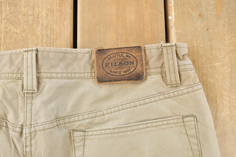 May include: Close-up of khaki-colored pants with visible stitching and a leather patch. The patch reads "SEATTLE, WA FILSON SINCE 1897". The pants have belt loops and a back pocket.
