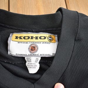 Puede incluir: Jersey de hockey negro con una etiqueta amarilla que dice "KOHO OFFICIAL LICENSED JERSEY" y "CHANDAIL LICENCIE OFFICIEL". La etiqueta tambi&eacute;n incluye informaci&oacute;n de tama&ntilde;o: "Adult, 2XL, 278, 209". El jersey est&aacute; hecho en Corea.