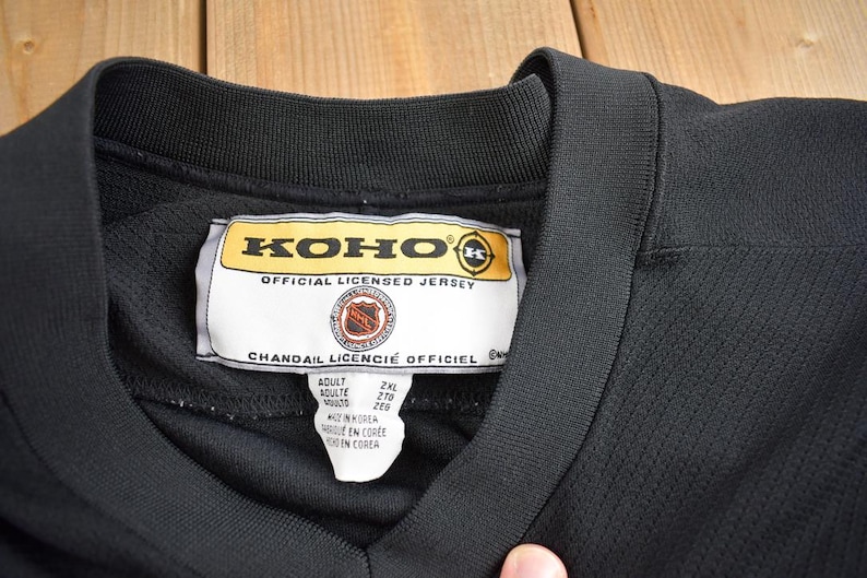 Puede incluir: Jersey de hockey negro con una etiqueta amarilla que dice "KOHO OFFICIAL LICENSED JERSEY" y "CHANDAIL LICENCIE OFFICIEL". La etiqueta tambi&eacute;n incluye informaci&oacute;n de tama&ntilde;o: "Adult, 2XL, 278, 209". El jersey est&aacute; hecho en Corea.