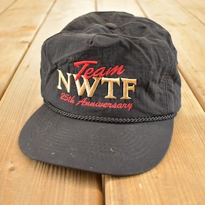 Op de afbeelding: Zwarte baseballcap met een gebogen klep. De cap heeft de tekst "Team NWTF 25th Anniversary" geborduurd in rode en gouden draad. Een zwart touwdetail accentueert de klep.