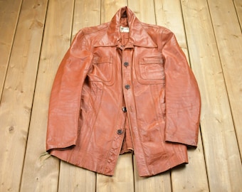 May include: A brown leather jacket with a button-up front and two flap pockets. The jacket has a collar and long sleeves.