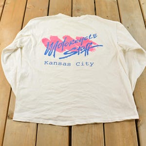 May include: White long-sleeve shirt with the text "MOTORCYCLE STAFF Kansas City" in blue and pink. The shirt has a crew neck and is made of a soft, comfortable material. The graphic is on the back of the shirt.
