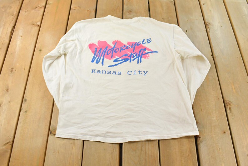 May include: White long-sleeve shirt with the text "MOTORCYCLE STAFF Kansas City" in blue and pink. The shirt has a crew neck and is made of a soft, comfortable material. The graphic is on the back of the shirt.