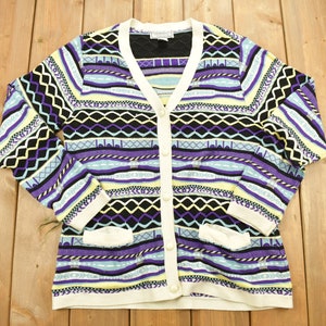 Puede incluir: Un cárdigan blanco con un estampado geométrico colorido en tonos de morado, azul, amarillo y negro. El cárdigan tiene cuello en V, mangas largas y cierre de botones.